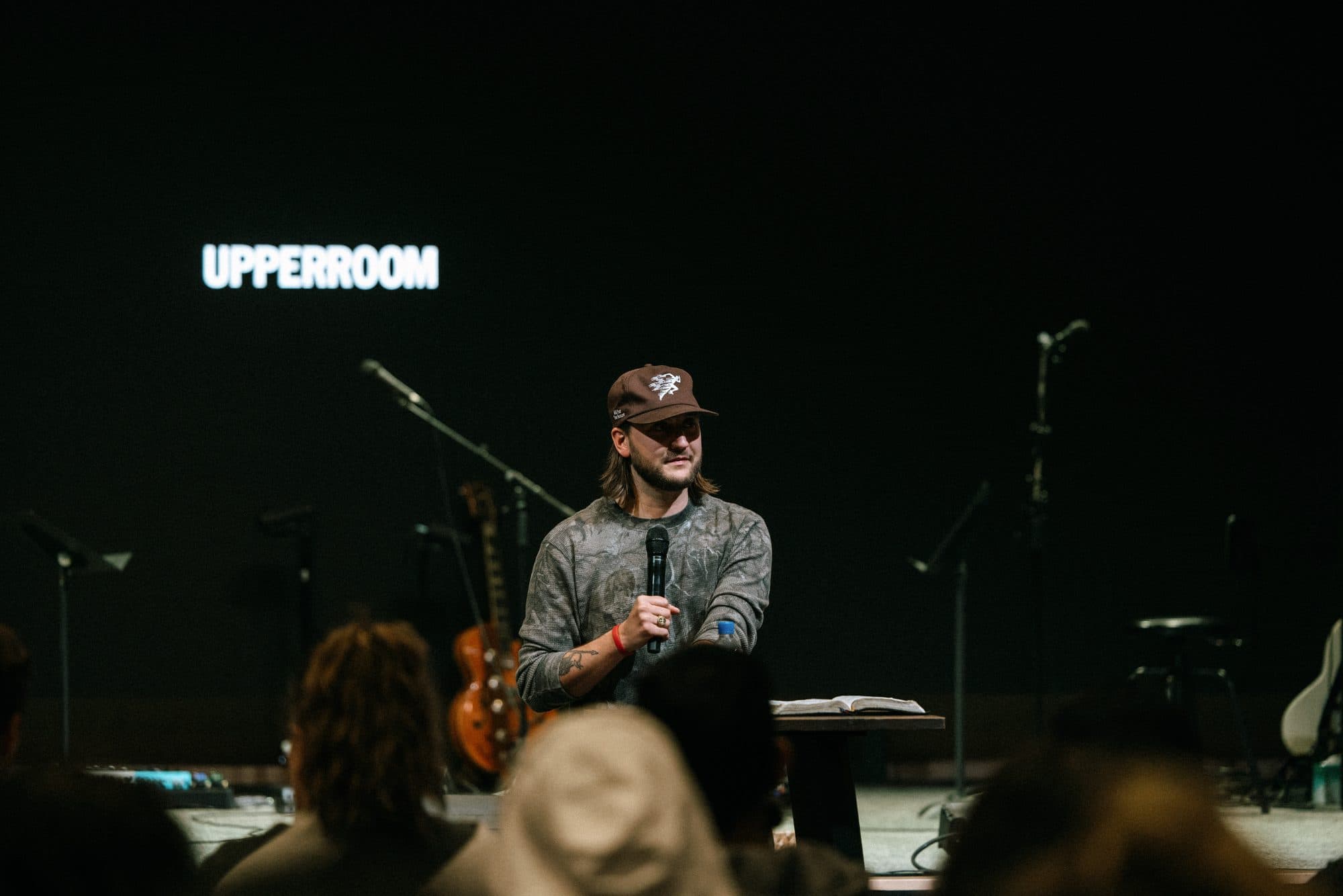 Chase preaching with a microphone on the UPPERROOM stage, the wordmark lit behind him